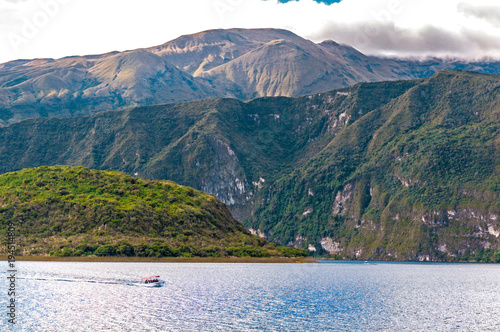 View of the Cuicocha lake and crater, with a small touristic boat in the water, on a sunny and cloudy afternoon. photo