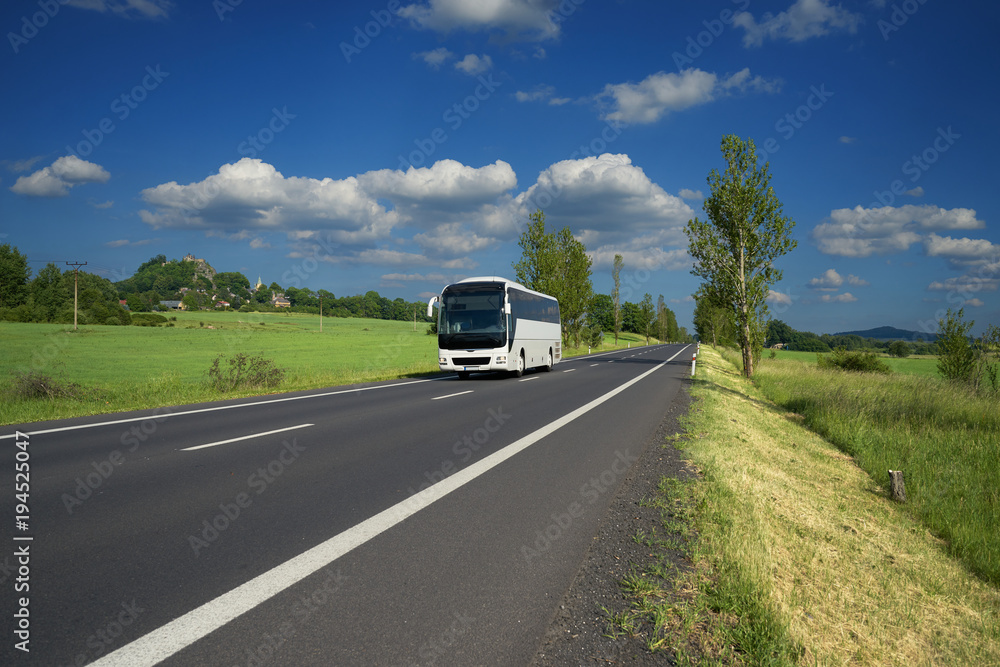 White bus traveling on asphalt road in a rural landscape. Village and mountain with castle ruins in the background.