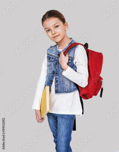 Beautiful little schoolgirl standing with rucksak and book isolated on grey background. Education, children and people concept photo