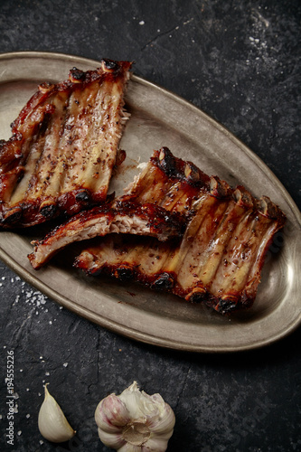 Closeup shot of flavorful grilled pork ribs in thick barbeque sauce cut in messy way served in vintage metal tray on dark textured background. Pub styling concept.
