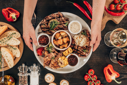 Φωτογραφία cropped image of woman putting plate with beef steaks, chicken wings and grilled