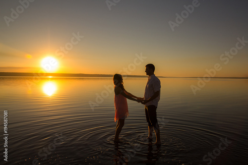 Young couple is holding hands in the water on summer beach. Sunset over the sea. . Two silhouettes against the sun. Calm and still surface of water. Romantic love story.