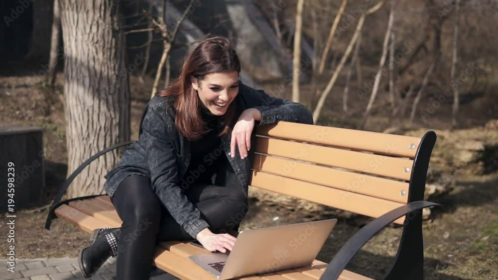Young attractive brunette girl is sitting on the bench in park waving at someone in her laptop, chatting, laughing and having fun time. Outdoors footage Young attractive brunette girl is sitting on the bench in park waving at someone in her laptop, chatting, laughing and having fun time. Outdoors footage