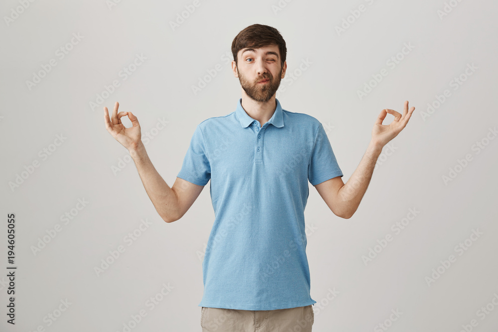 Funny attractive caucasian man with beard and moustache peeking and making faces while standing in meditating pose with zen gesture as if he is doing yoga over gray background Funny attractive caucasian man with beard and moustache peeking and making faces while standing in meditating pose with zen gesture as if he is doing yoga over gray background