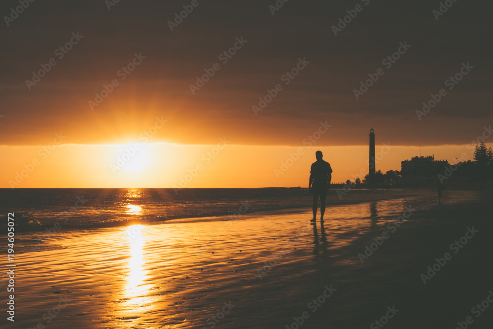 Walker on seashore with lighthouse during sunset in Maspalomas, Gran Canaria Island.