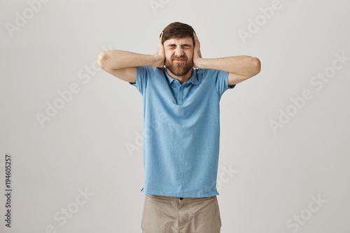 Noise is killing me. Studio shot of displeased irritated caucasian male covering ears with palms while squinting and being disgusted with sound he hears, standing over gray background. Turn off alarm photo