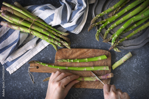 Fresh  raw asparagus on stone background. Women female hands cut asparagus on wooden cooking board. Top view