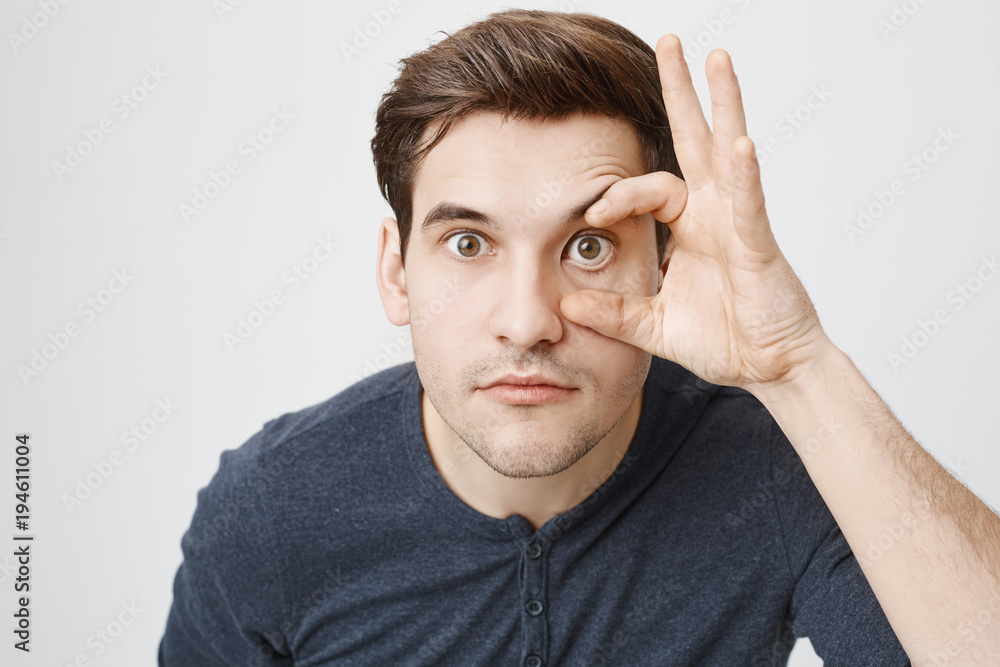 Shot of funny positive dark-haired guy grimacing, making faces, looking carefully at camera. Man with stubble has serious expression of face while posig indoors. Body language and face expression Shot of funny positive dark-haired guy grimacing, making faces, looking carefully at camera. Man with stubble has serious expression of face while posig indoors. Body language and face expression