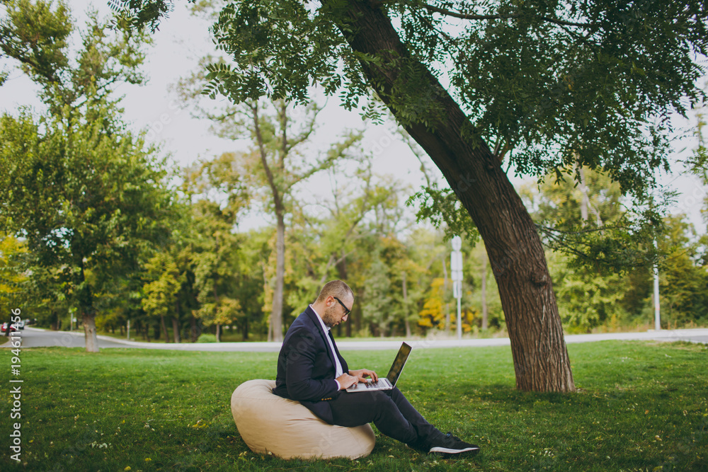 Young successful businessman in white shirt, classic suit, glasses. Man sit on soft pouf, working on laptop pc computer in city park on green lawn outdoors on nature. Mobile Office, business concept. Young successful businessman in white shirt, classic suit, glasses. Man sit on soft pouf, working on laptop pc computer in city park on green lawn outdoors on nature. Mobile Office, business concept.