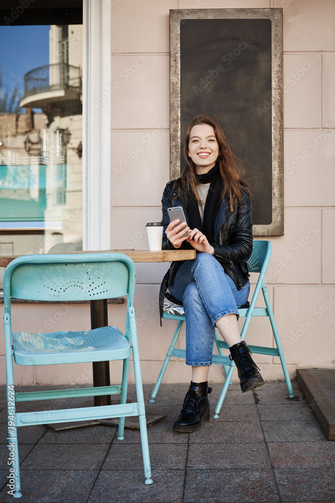 There is always time for coffee. Portrait of happy good-looking woman sitting in patio of cafe, holding smartphone and looking with broad smile at camera, drinking tea while waiting for her boyfriend