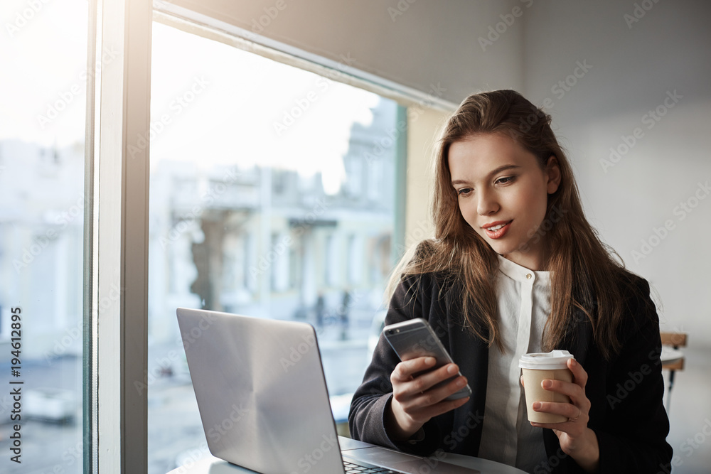 Fototapeta premium Surrounded with gadgets. Indoor portrait of attractive caucasian businesswoman sitting in cafe near window, drinking coffee and messaging via smartphone, working with laptop during break