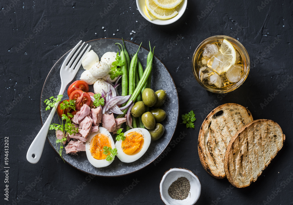 Aperitive table - plate of canned tuna, green beans, mozzarella cheese, tomatoes, boiled egg, olives, grilled bread and whiskey glasses on dark background, top view Aperitive table - plate of canned tuna, green beans, mozzarella cheese, tomatoes, boiled egg, olives, grilled bread and whiskey glasses on dark background, top view