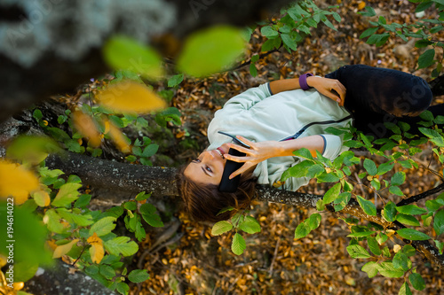 Young woman lying on a branch, in a tree, talking on her smart phone, smiling and feeling relaxed in nature in autumn season