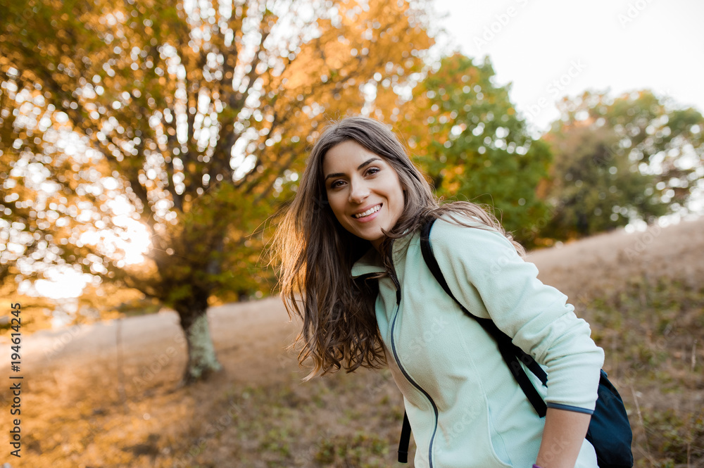Young beautiful woman on a hill in the forest with backpack in the autumn season, at sunset, looking happy and smiling 