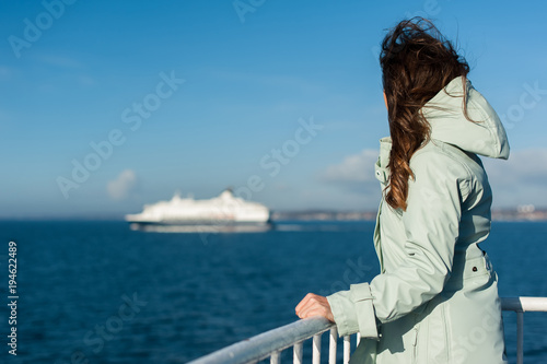 Stampa su Tela Young traveller woman sailing a ferry, with big boat cruise liner or ferry on the background, wearing a rain jacket