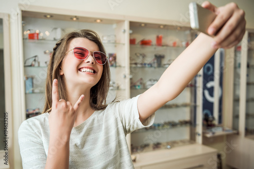 This optician shop rocks. Portrait of attractive female blogger in store trying on trendy sunglasses and showing rock gesture, smiling broadly, being happy to find perfect glasses for everyday usage