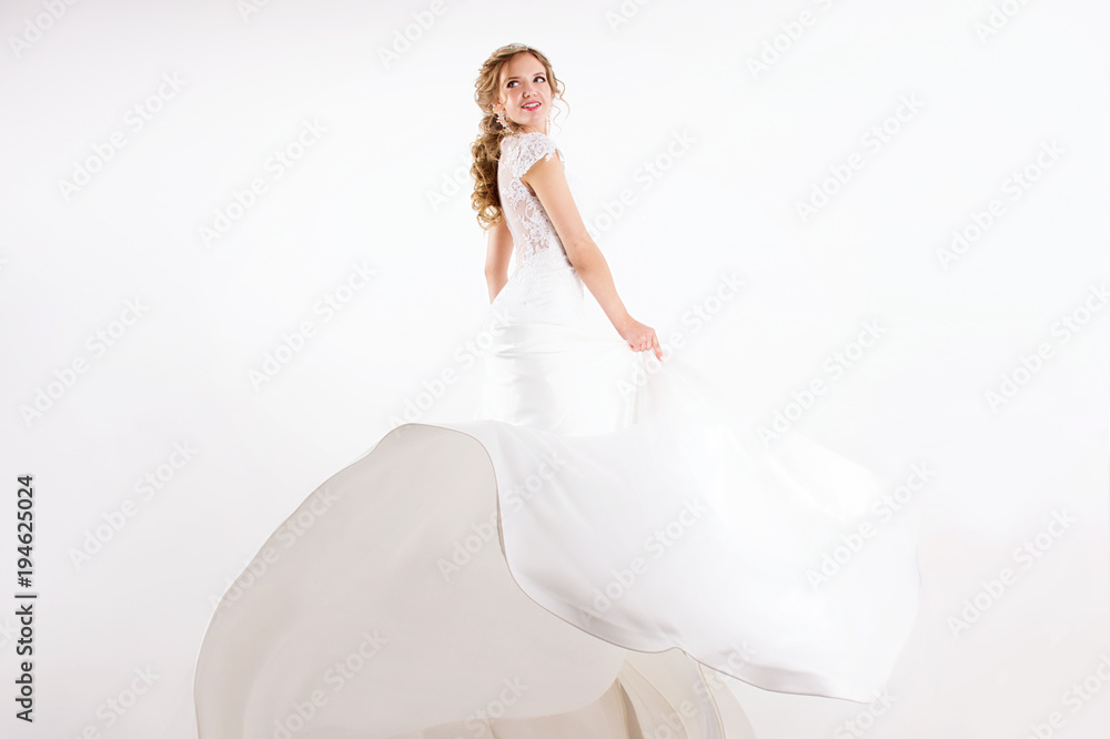 young blonde girl in a white wedding dress and jewelry in hairstyle  with big curls standing like princess and smiling on a white wall background 