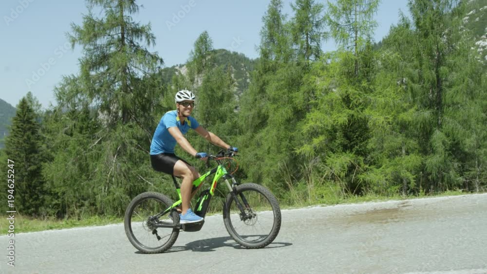 SLOW MOTION: Cheerful cyclist riding his innovative electric bike up a hill in the sunny mountains. Happy Caucasian male rides cool e-bike and looks at beautiful green and sunny nature surrounding him