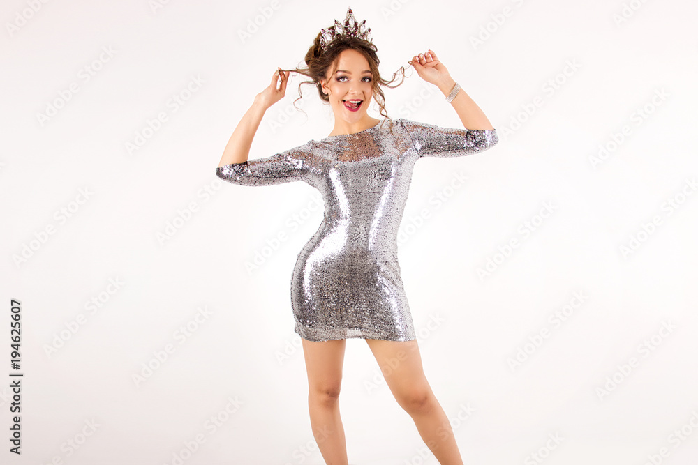 young girl in a silver shiny disco dress with curls and big crown standing and smiling, crooked and showing tongue on a white wall background young girl in a silver shiny disco dress with curls and big crown standing and smiling, crooked and showing tongue on a white wall background