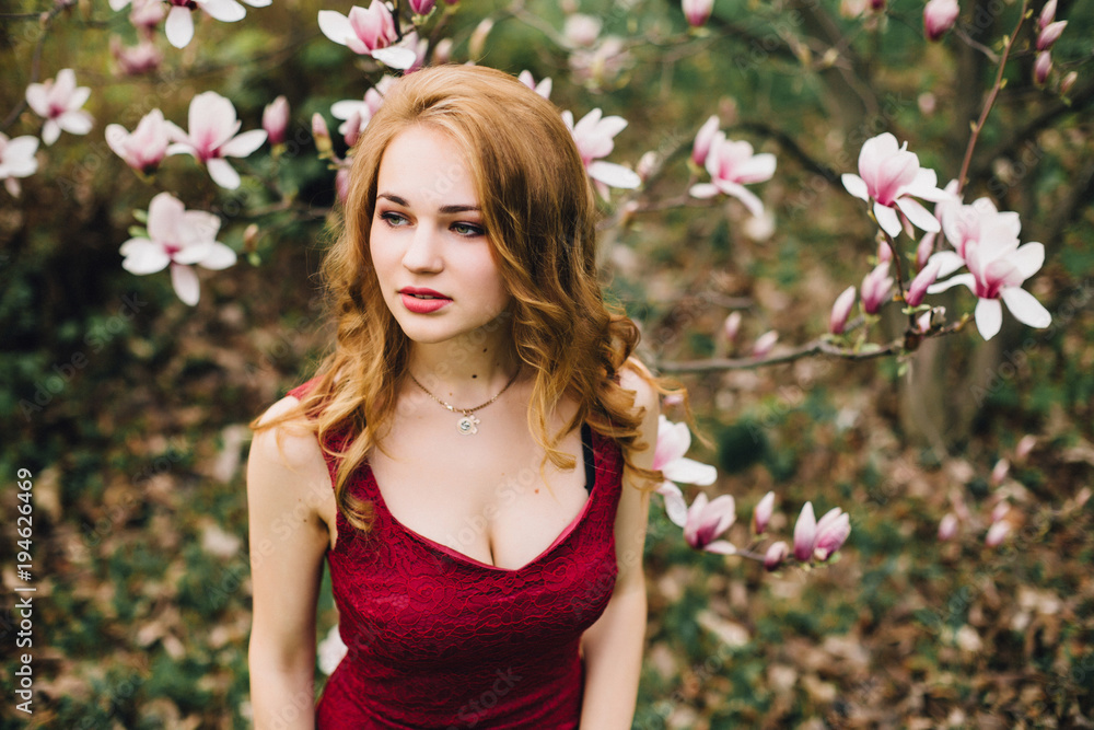 Fototapeta premium Beautiful girl in the flowering cherry blossom. In a burgundy dress with beautiful hair and makeup