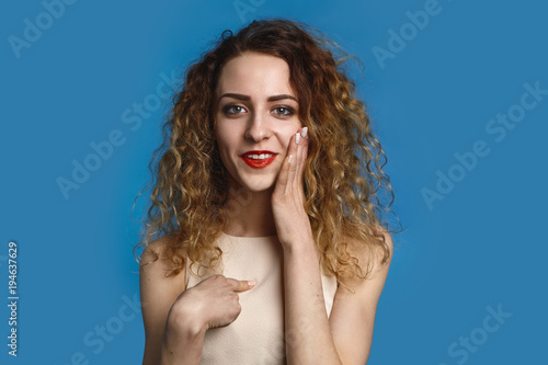 Portrait of emotional positive young Caucasian woman with loose curly hair posing in studio, having excited look, smiling happily at camera, touching face and pointing index finger at herself