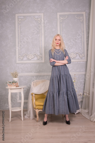 The blonde girl in a long gray dress crossed her arms on her chest and stands in vintage interiors