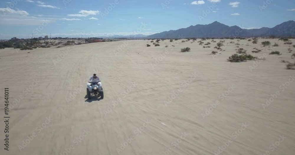 Aerial camera moving backward rapidly as an ATV comes toward it, bouncing over tracks in the sand with mountains in the distance. Aerial camera moving backward rapidly as an ATV comes toward it, bouncing over tracks in the sand with mountains in the distance.