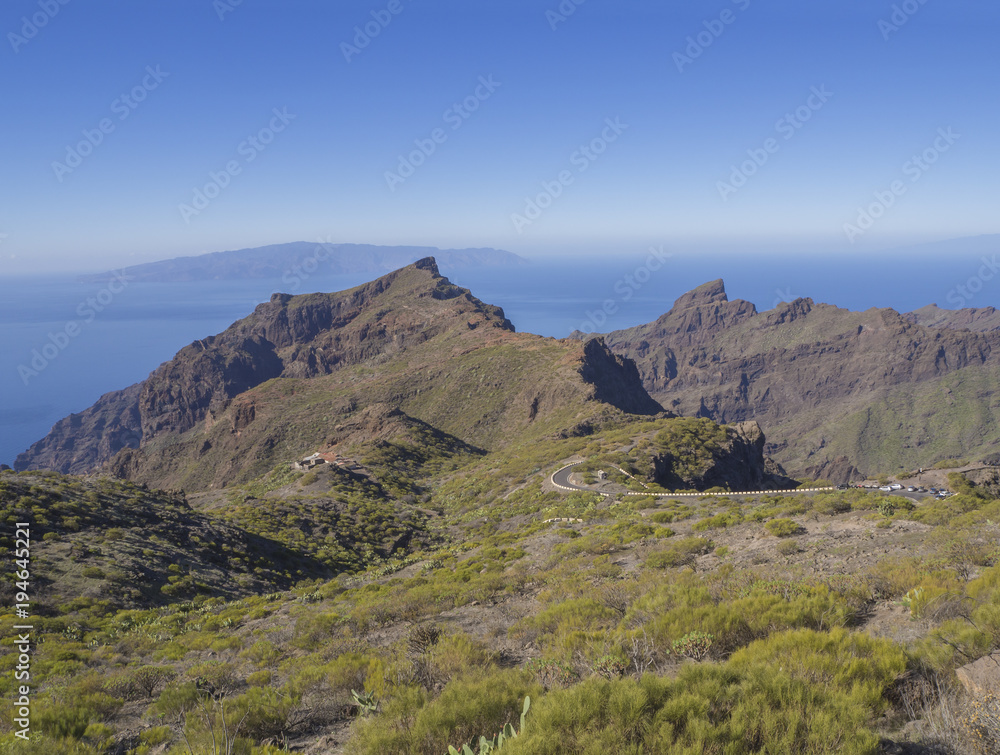 view on asfalt road to village Masca with green hills, sharp mountain peaks, sea horizon and clear blue sky background view on asfalt road to village Masca with green hills, sharp mountain peaks, sea horizon and clear blue sky background