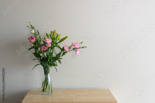 Pink lisianthus flowers and green foliage in glass vase on wooden shelf against beige wall