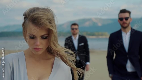 Blonde girl is going out from two handsome men on a river coast. She can not choosing between them, love triangle
