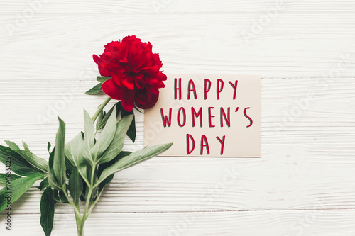 happy women's day text sign on craft card and beautiful red peonies on white wooden rustic background, flat lay. modern greeting card. womens day