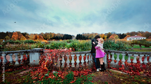 Love story of young beautiful couple in Peterhof, Saint-Petersburg, Russia at autumn. Blonde girl in pink coat and blue dress and man or groom in black jacket and jeans. Embrace kiss with fall in love photo