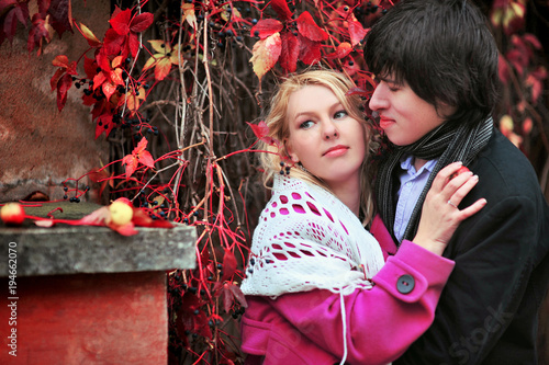 Love story of young beautiful couple on the red leaves background at autumn. Blonde girl in pink coat and blue dress and man or groom in black jacket and jeans. Embrace kiss with fall in love photo