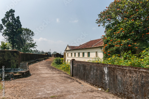 Madikeri, India - October 31, 2013: Walkway and defensive wall on top of ramparts of fort. Green vegetation. Gray wall. Blue sky. Yellow building on side.