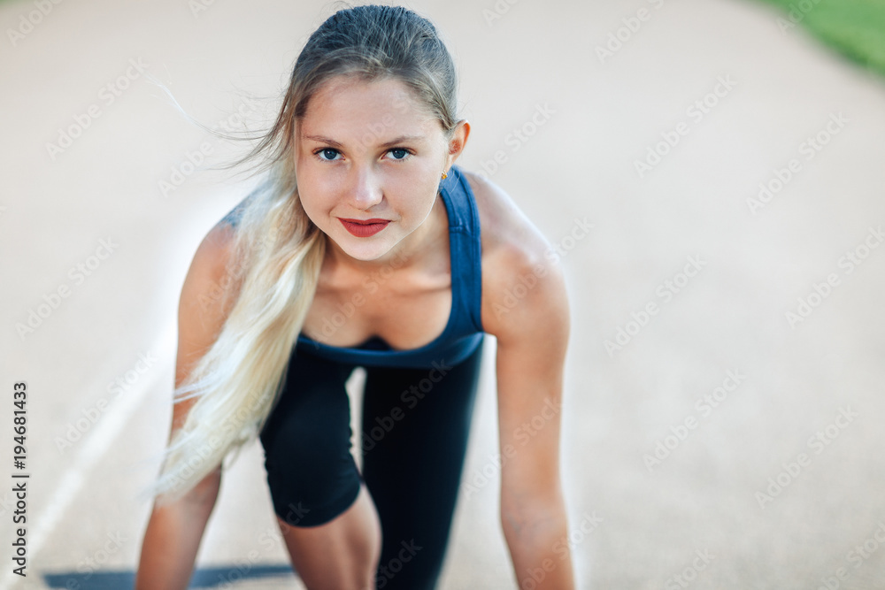 Ready to go! Female athlete in a blue shirt and leggings on the starting line of a stadium track looking at camera and smiling, preparing for a run. Fitness girl is preparing to run from a low start. Ready to go! Female athlete in a blue shirt and leggings on the starting line of a stadium track looking at camera and smiling, preparing for a run. Fitness girl is preparing to run from a low start.
