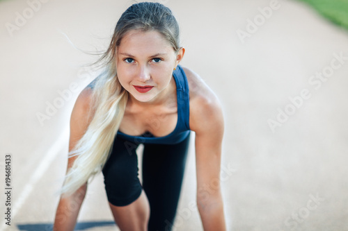 Ready to go! Female athlete in a blue shirt and leggings on the starting line of a stadium track looking at camera and smiling, preparing for a run. Fitness girl is preparing to run from a low start.