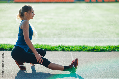 Fitness girl in a blue shirt and leggings doing warm up routine on the sadium before training, stretching body muscles. Female athlete preparing legs for cardio workout. photo