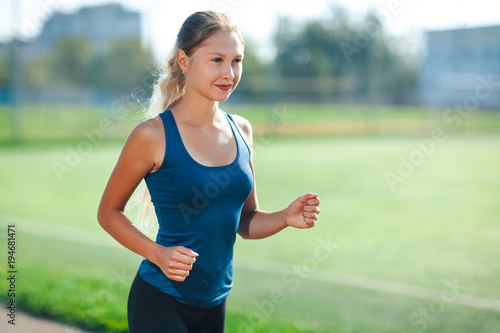 Young fitness woman in a blue shirt and leggings running on a stadium track. Athlete girl doing exercises on the training at stadium. Healthy active lifestyle.
