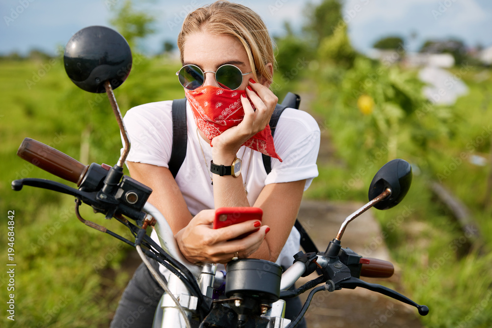 Fashionable female driver dressed casually, reads bikers blog on mobile phone, sits on motorbike, freshes fresh air outdoor, looks thoughtfully into distance. People, lifestyle and technology