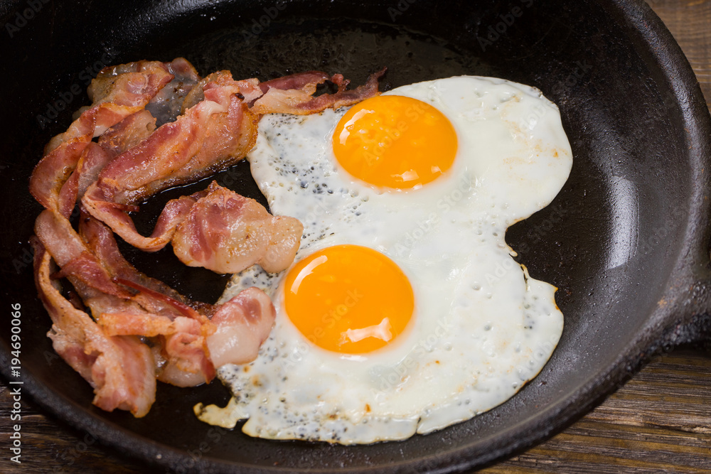 Bacon and Egg. Salted egg and sprinkled with black pepper. English breakfast. Grilled bacon, two eggs on pan on wooden table. Bacon and Egg. Salted egg and sprinkled with black pepper. English breakfast. Grilled bacon, two eggs on pan on wooden table.