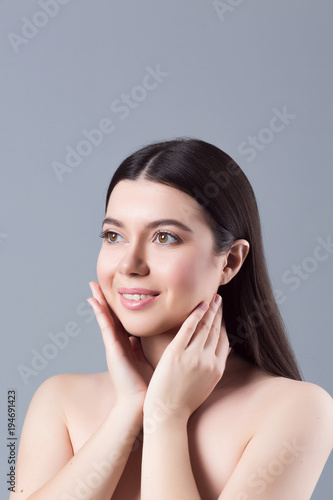 Beautiful young woman with clean skin isolated on grey background. Portrait of beauty girl with natural nude makeup touching her face. Spa, skincare and wellness concept close up.