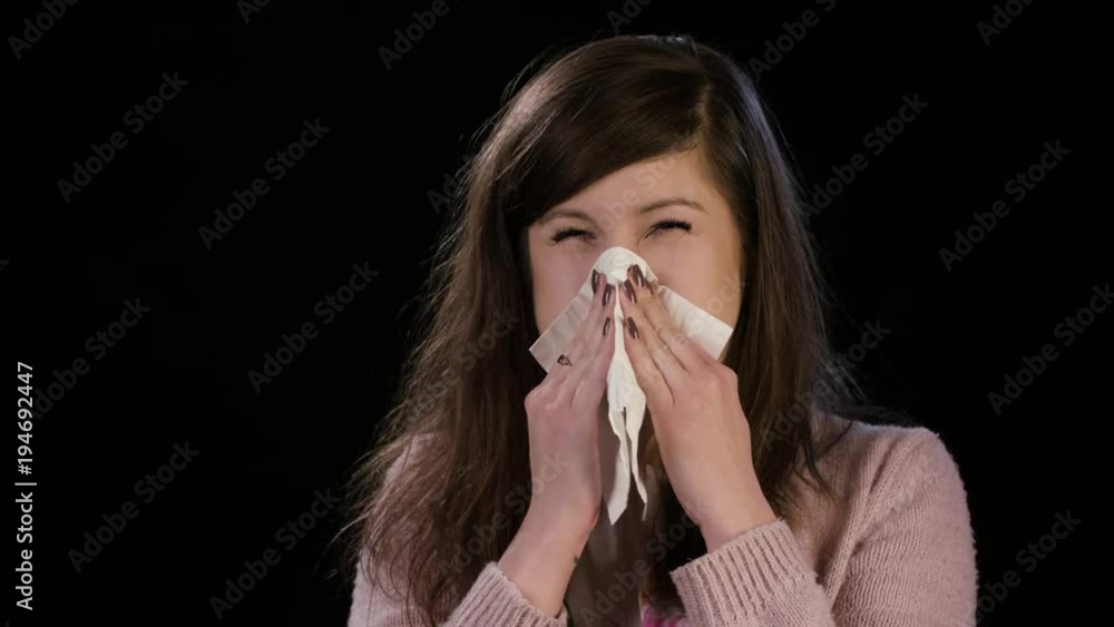 An attractive sick young lady wearing a pink sweater clearing her nose against a black background. Close-up An attractive sick young lady wearing a pink sweater clearing her nose against a black background. Close-up