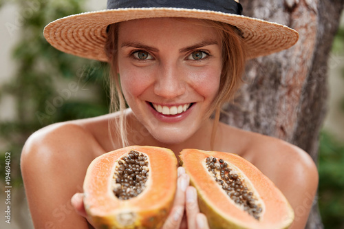 Horizontal portrait of glad lovely woman poses naked, wears summer hat, holds organic exotic papaya, supports healthy eatting. Beautiful female likes tropical fruits, spends summer at hot country. photo