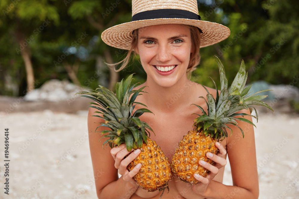 People, fitness, healthy eating concept. Adorable woman has positive  expression, hides her naked body with two pineapples, spends free time at  beach, sunbathes alone. Female with fresh fruit foto de Stock