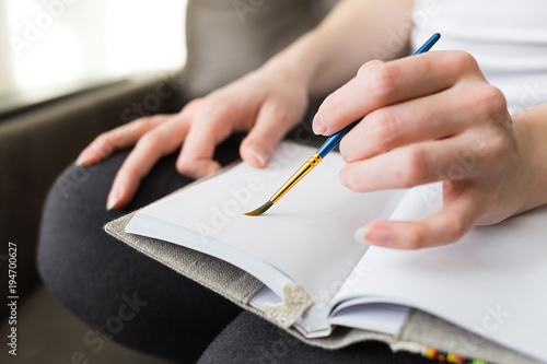 A girl holding a brush in her hands and drawing in a notebook that lies on her lap close-up.