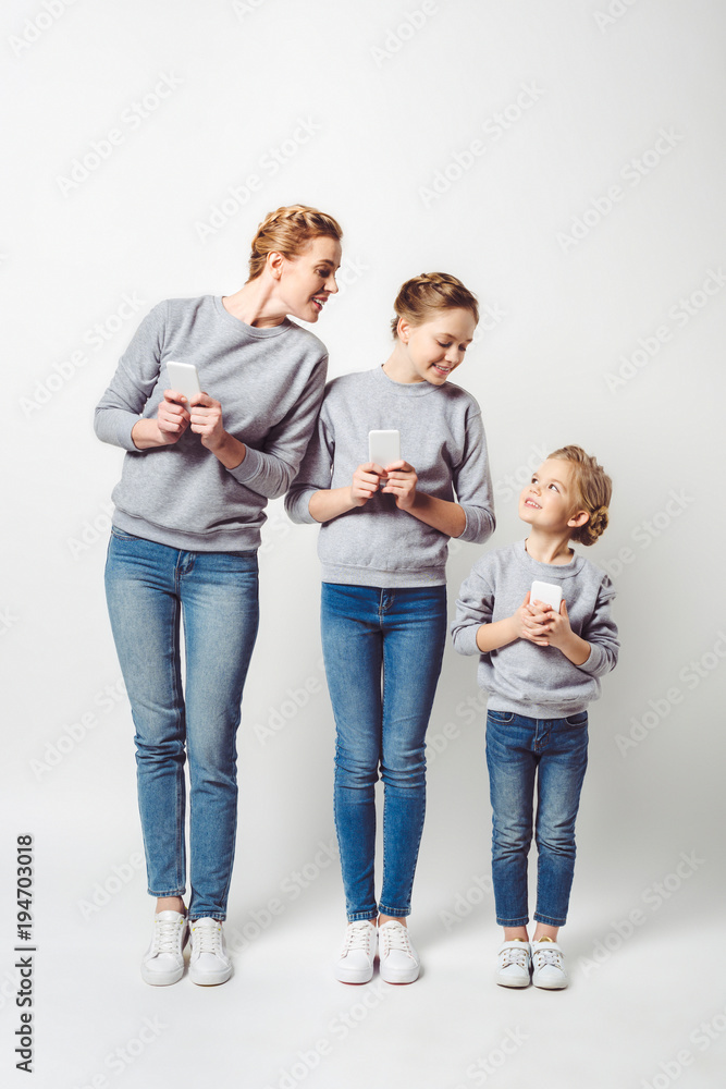 smiling mother and daughters in similar clothing with smartphones isolated on grey smiling mother and daughters in similar clothing with smartphones isolated on grey