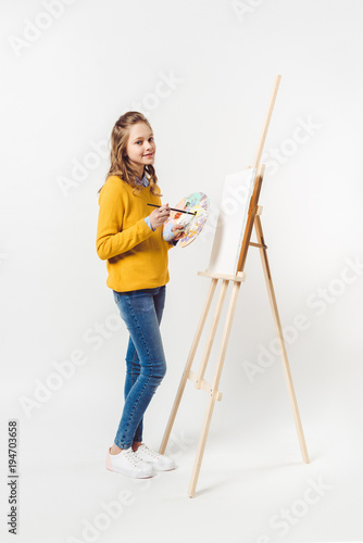 teen female painter with paint brush and palette near easel with blank canvas on white