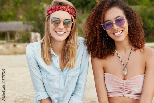 Mixed race young female models with happy expressions  sit together at tropical beach  wear sunglasses  happy to meet  demonstrate true friendship or homosexual relationships. Feminity concept