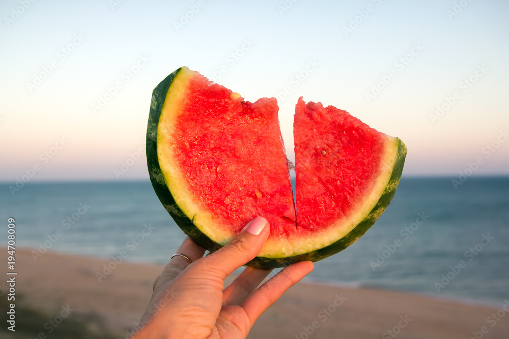 Juicy watermelon slice in the hand of a young girl on the background of the sea coast, symbol of summer Juicy watermelon slice in the hand of a young girl on the background of the sea coast, symbol of summer