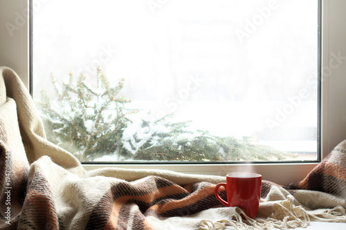 cozy home atmosphere in the winter/ red mug of hot drink on a windowsill with a warm blanket, when behind is window is snow